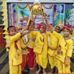 Children in yellow traditional outfits raise a decorated ceremonial pot on sticks during a festival procession